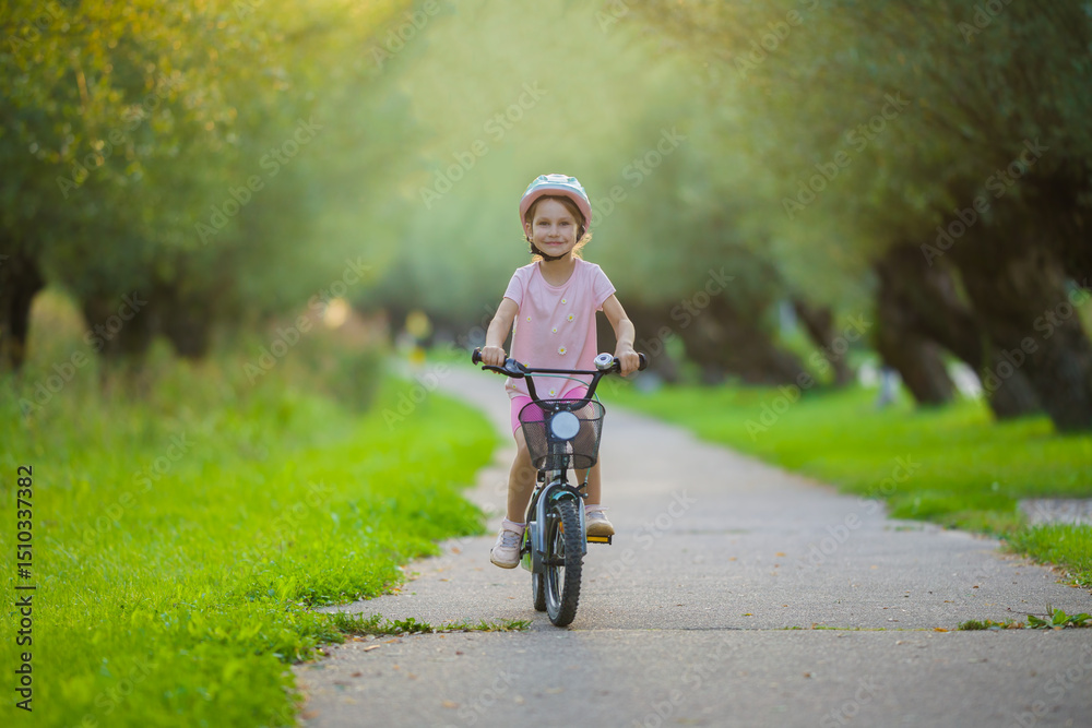 Fototapeta premium Happy smiling little child girl with helmet riding on bike on road at town park in warm summer day. Enjoying cycling. Cute 5 years old toddler. Front view.