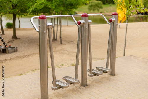 Fototapeta Naklejka Na Ścianę i Meble -  A set of outdoor fitness equipment designed for leg workouts is positioned alongside a peaceful park setting with a calm water body in the background, surrounded by trees