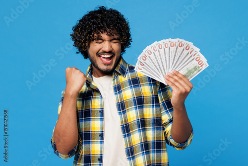 Young happy Indian man he wearing yellow checkered shirt casual clothes hold in hand fan of cash money in dollar banknotes do winner gesture isolated on plain blue cyan background. Lifestyle concept.