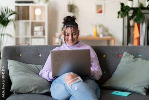 Smiling young woman sits on a comfortable couch at home, using her laptop for work or leisure. She is relaxed and enjoying her time online.