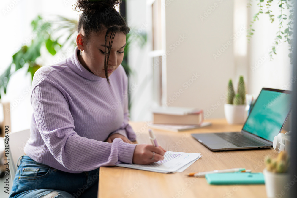 Young woman studying at a desk, writing on papers with a pen. Laptop and books are nearby. She is focused on her work in a bright room. Young woman studying at a desk, writing on papers with a pen. Laptop and books are nearby. She is focused on her work in a bright room.