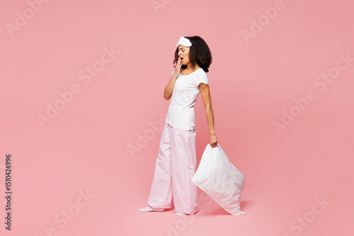 Full body calm young woman wear pyjamas jam sleep eye mask rest relax at home stand hold pillow yawn cover mouth with hand isolated on plain pink background studio portrait Good mood night nap concept