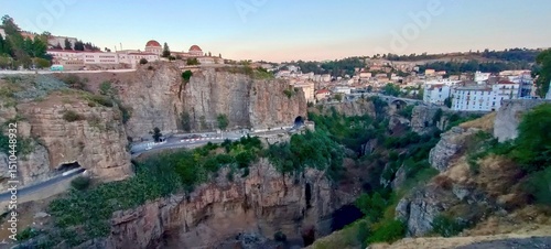Dramatic landscape of Constantine, Algeria, during the golden hour light. It showcases the deep gorges carved by the Rhumel River, iconic limestone cliffs, the Bab Al-Qantara Bridge in the distance.