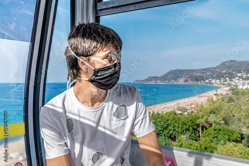 Man in a face mask enjoying the view of the Mediterranean Sea from the Alanya Teleferik cable car, Turkey. The background showcases a coastal landscape with blue waters and distant mountains