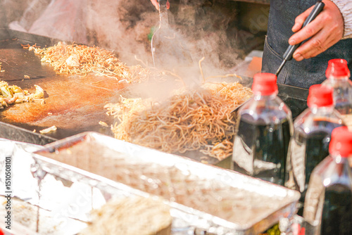 Steaming yakisoba noodles and vegetables sizzle on a large teppanyaki grill, expertly prepared by a vendor at a bustling street food stall