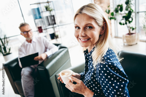 Smiling mature businesswoman in stylish attire holding coffee in modern office interior with male colleague in background Free Download Smiling mature businesswoman in stylish attire holding coffee in modern office interior with male colleague in background - GFXLoad