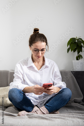 Young woman in glasses in a white shirt looks at the phone while sitting on the sofa.