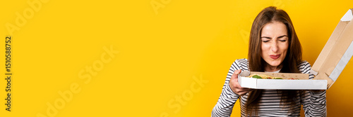 cheerful young woman enjoying the aroma of fresh hot pizza on yellow background. Banner