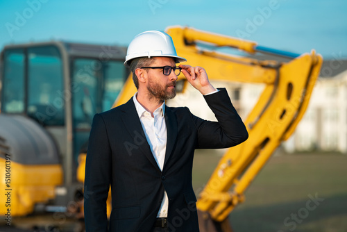 Architect at a construction site. Architect man in helmet and suit at modern home building construction. Architect with a safety vest and suit. Confident architect standing at house background.