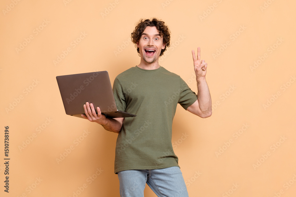 Happy young man with curly hair holding a laptop and gesturing a peace sign in casual style on a beige background Happy young man with curly hair holding a laptop and gesturing a peace sign in casual style on a beige background
