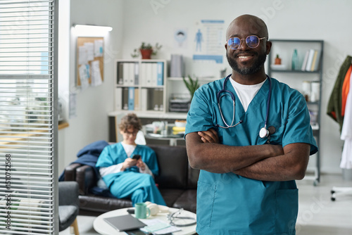 Wallpaper Mural African American doctor standing with arms crossed, smiling confidently in modern medical office, while a nurse sits on couch in background, busy with paperwork Torontodigital.ca