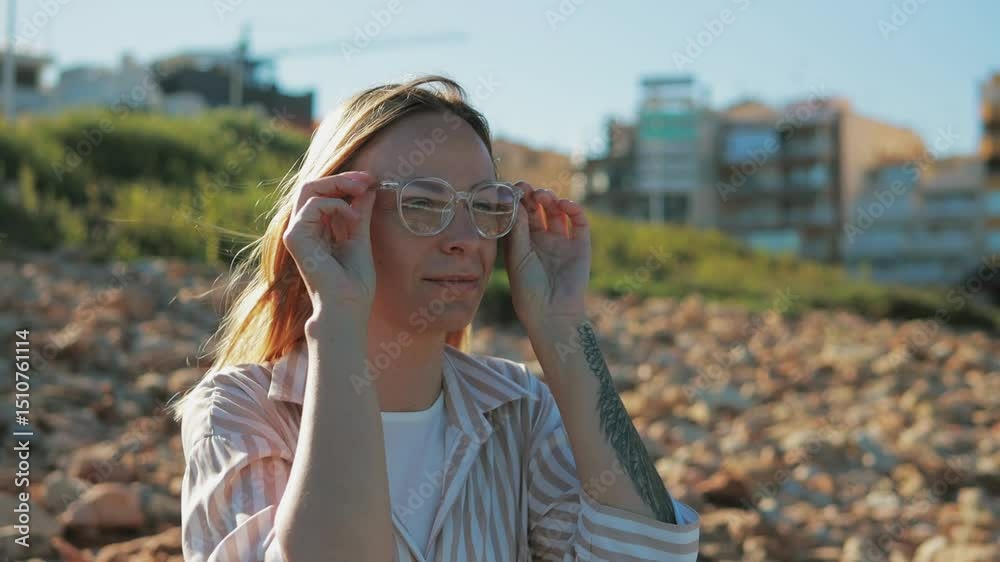 Blonde woman in striped shirt standing on rocky coastline, adjusting transparent glasses while gazing at serene ocean landscape under sunlight Blonde woman in striped shirt standing on rocky coastline, adjusting transparent glasses while gazing at serene ocean landscape under sunlight