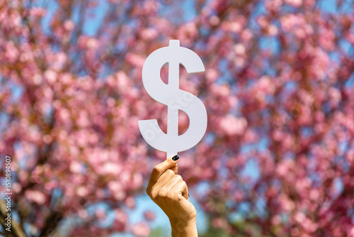 Hand holding a white dollar sign symbol in front of blooming pink cherry blossoms under a clear blue sky.