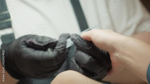 Nail technician wearing black gloves precisely filing customer's fingernail using hand filer during manicure session, both hands clearly visible in close up with customer receiving nail care