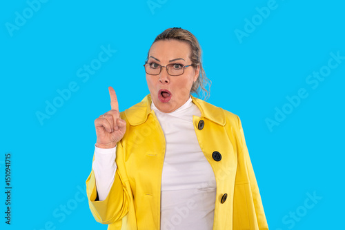 Woman in yellow coat gestures emphatically while standing against a bright blue background in a lively expression of surprise or concern photo
