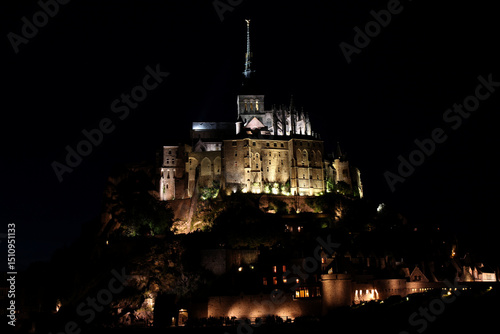 Fototapeta Naklejka Na Ścianę i Meble -  Night scene of Mont St Michel in France, with buildings lit by street lights and the tower lit by a search light, Normandy, northern France