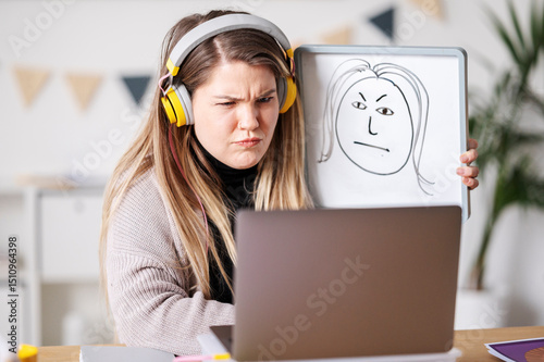 A woman with headphones is video conferencing, looking displeased. She's holding up a whiteboard with a drawing of a face that mirrors her expression, perhaps to illustrate her feelings. photo