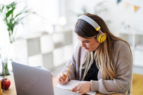 A student is studying at home. She's wearing headphones and taking notes from her laptop to prepare for an upcoming exam.