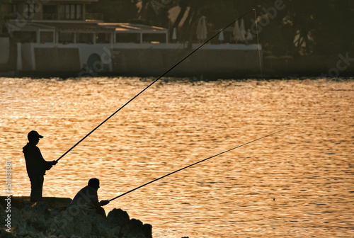 Silhouettes of two fishermen standing by a calm waterfront, casting their fishing rods during a serene sunset. The golden hues reflect peacefully on the water, embodying tranquility and patience.