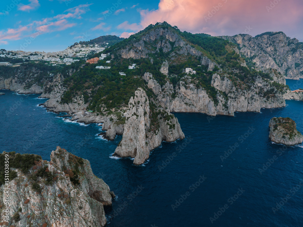 Fototapeta premium Capri Island's coastline features rocky cliffs, jagged sea stacks, lush greenery, and white hillside buildings under a pink and blue sky.