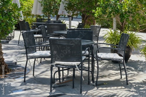 Cozy outdoor seating area of empty restaurant or bistro surrounded plant on flower pots. Black tables and chairs with pads on patio terrace or cafes.