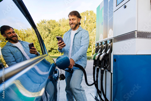 Quadro su tela Happy handsome man refueling his car and messaging on smartphone, filling up aut