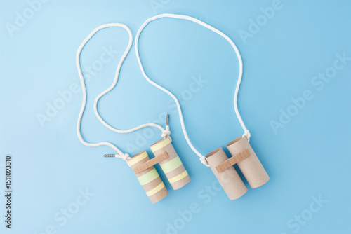 Two pairs of handmade toy binoculars made from cardboard toilet paper rolls and white string on a blue background. Flat lay composition. DIY kids craft and recycling education concept.