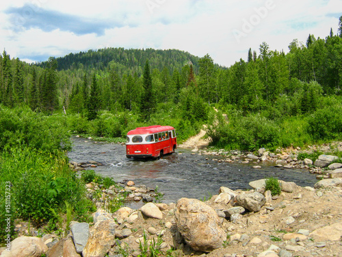 A red bus crosses a shallow mountain river, transporting supplies and hikers in the green wilderness of the Kuznetsk Alatau Mountains. photo