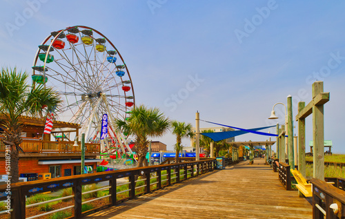 A sunny view down a wooden boardwalk and a large Ferris wheel at the Carolina Beach Amusement Park in Wilmington, North Carolina in the USA.