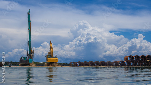 excavator and crane, large diameter metal casing pipes stacked. Beautiful background of blue sky and sea