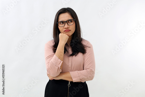Young Asian woman standing with folded arms and supporting chin with hand, annoyed, angry, thinking expression, wearing formal shirt, glancing sideways into empty space, isolated on white background.
