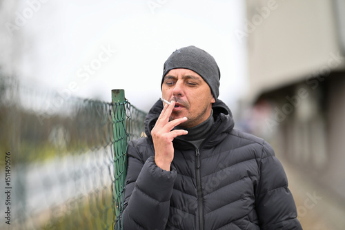 Man Smoking Outside.Man in winter jacket and beanie smoking a cigarette near a chain link fence outdoors.
