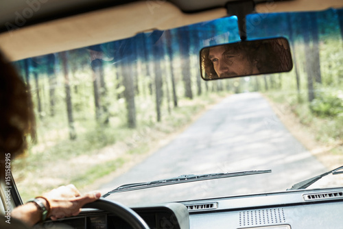 Hands gripping steering wheel while mirror shows reflection of face, with lush forest visible through windshield and rearview mirror, creating serene driving experience photo