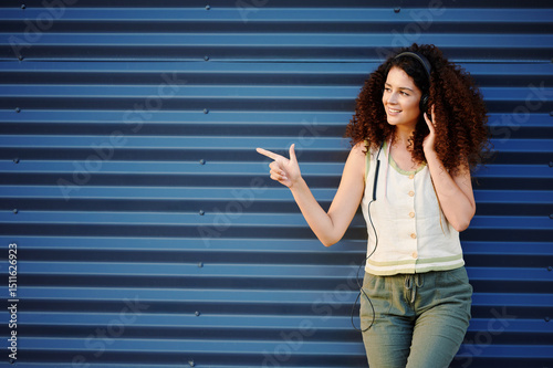 woman enjoying music through headphones against blue wall. casual style with curly hair, green pants, sleeveless top. pointing hand gesture, relaxed expression, outdoor urban scene photo