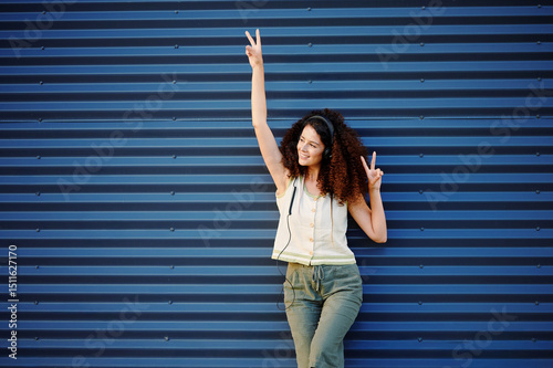 confident woman with headphones enjoys music while standing against blue metal wall, expressing joy with peace signs. casual style, outdoor setting evokes freedom and fun photo