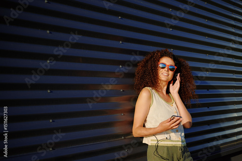 woman with smartphone enjoying sunny day. young woman with curly hair wearing sunglasses, listening to music outdoors against modern wall. urban lifestyle and relaxation concept photo
