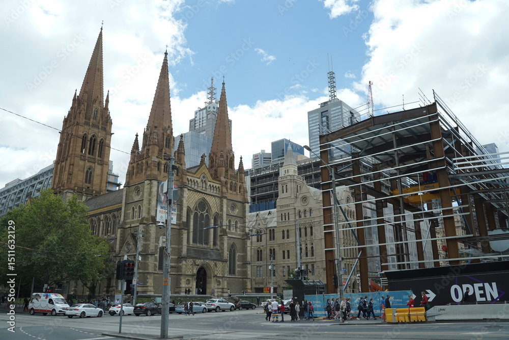 Naklejka premium A wide shot captures a historic cathedral with two prominent spires standing beside a modern building under construction, with a city skyline visible in the background under a cloudy sky. Cars and ped