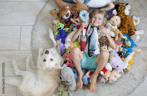 barefoot child lies on the floor among a pile of various colorful soft toys, next to the pet - a white dog. Play at home with toys, be friends with animals. Develop imagination, interesting childhood