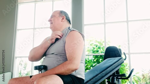 Senior overweight man experiencing chest discomfort while exercising at the gym, touching his chest in pain. Concept for heart health, medical awareness, and workout risk in elderly fitness.
