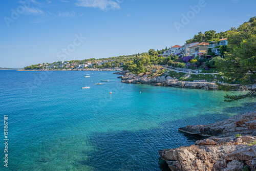 Fototapeta Naklejka Na Ścianę i Meble -  A tranquil Mediterranean beach with clear blue water, pine trees, rocky shoreline, and distant islands under a sunny sky.