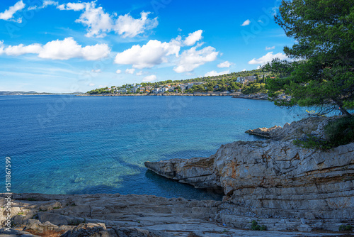 Fototapeta Naklejka Na Ścianę i Meble -  A tranquil Mediterranean beach with clear blue water, pine trees, rocky shoreline, and distant islands under a sunny sky.