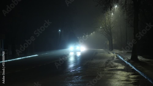 Headlights pierce the fog as cars move on an eerie, dimly lit street.