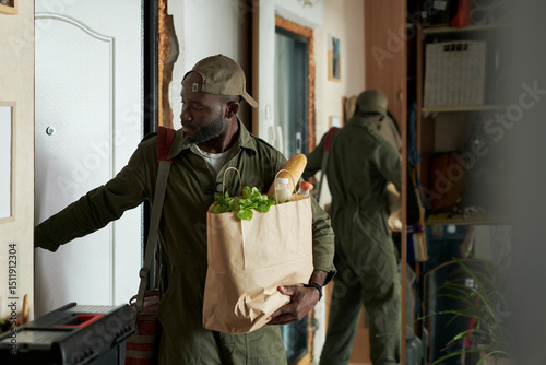 Man carrying groceries entering front door with reflection visible in mirror. Holding a paper bag filled with fresh produce and bread, fully focused on opening the door