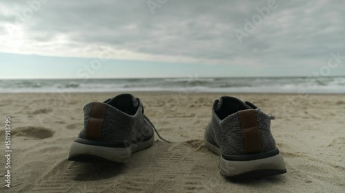 Abandoned shoes on sandy beach with waves in background, cloudy sky overhead.