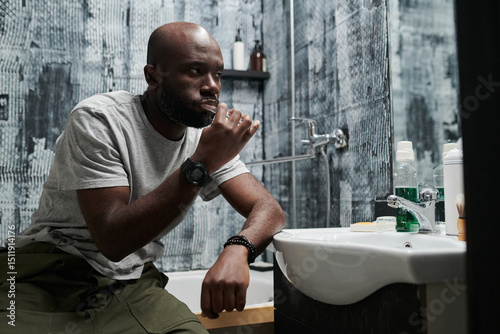 Man with bald head and beard sitting in bathroom brushing teeth, lost in thought and contemplation, wearing a casual shirt and pants, modern bathroom setting with toiletries visible