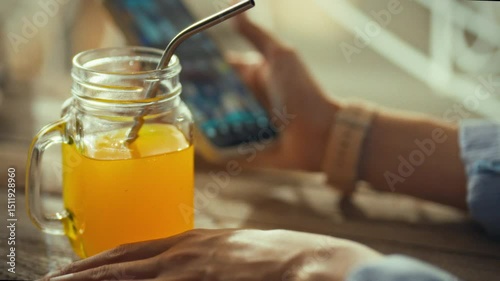 A person is relaxing in a cozy cafe, sipping a glass of bright orange juice through a metal straw while checking their smartphone. The atmosphere is inviting and casual