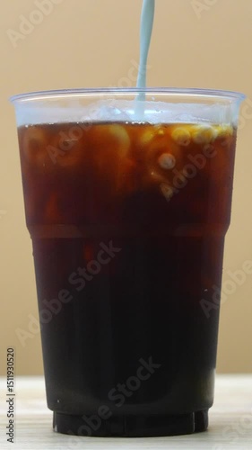 Vertical video Close-up iced black coffee in a plastic cup stream of milk is poured in, creating a dramatic swirl effect.