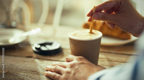 A hand stirs a cup of warm coffee at a rustic table in a cozy cafe. Sunlight filters through the window, highlighting a croissant nearby, perfect for a leisurely morning