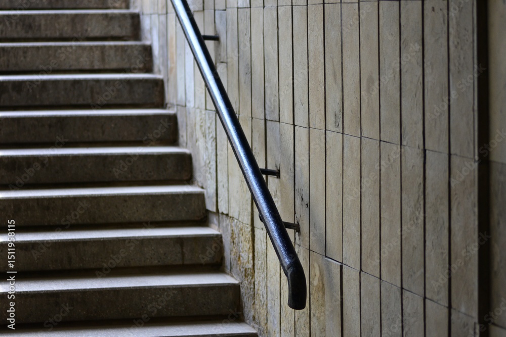 Obraz premium Close-up view of a black metal handrail on a tiled wall beside concrete stairs in an indoor public space. Ideal for themes of architecture, safety, structure, and modern urban design.