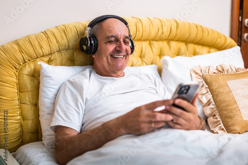 Cheerful senior man in pajamas using wireless headphones and mobile phone while comfortably sitting on bed in his bedroom.